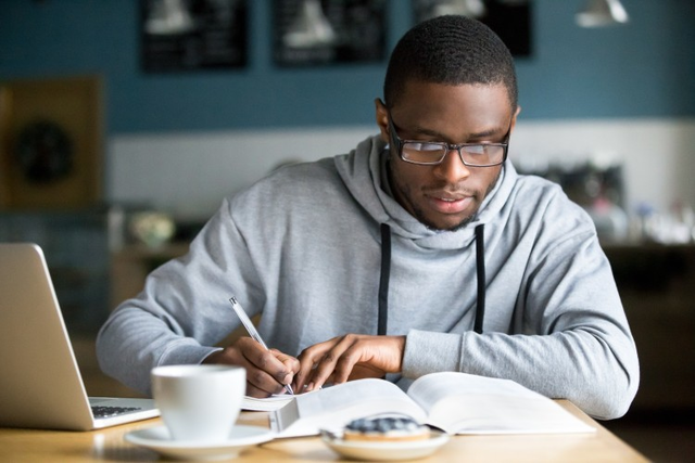 A young man takes notes as he reads a textbook at his kitchen table.