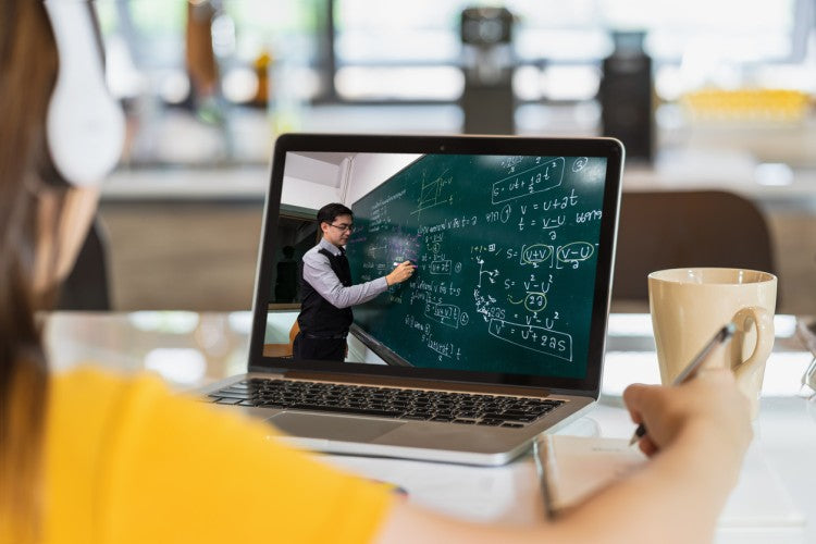 A high school student sits at a laptop watching a video of a math teacher writing equations on a blackboard.