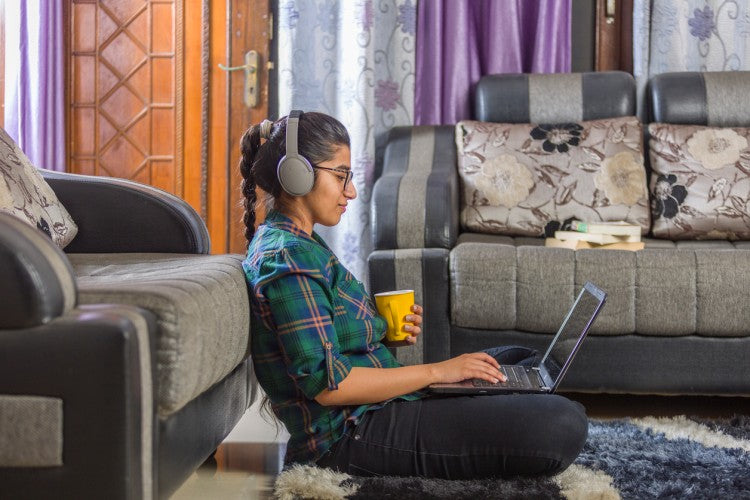 A high school student wearing headphones sits cross-legged on the floor in her living room holding a laptop and a coffee cup.