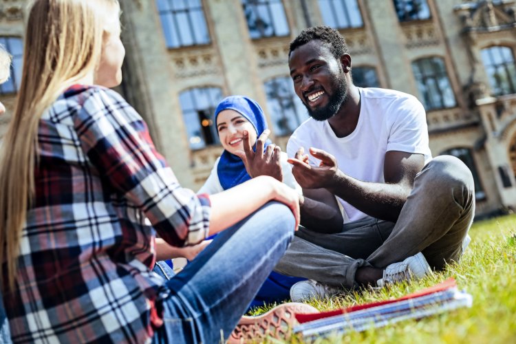 Three students in a study group sit on the lawn of a school campus talking. 