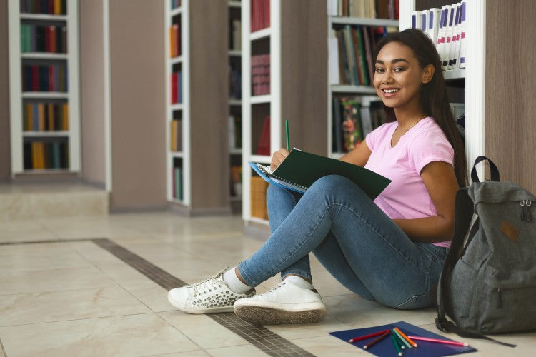 A high school student sits in the library writing in a notebook.