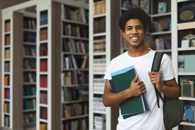 A student wearing a backpack and holding books stands in a school library. 