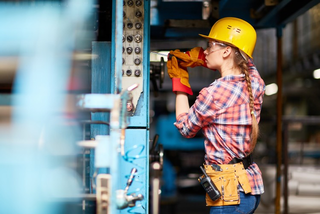 Electrician wearing hardhat and toolbelt repairs an industrial machine.